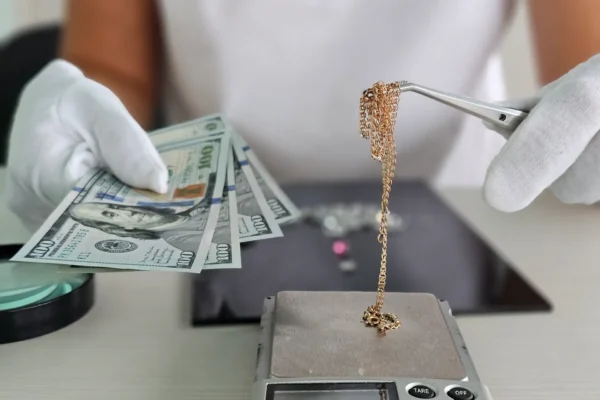 A jeweler weighs a gold chain on a precision scale while holding several banknotes in his other hand in a jewelry store