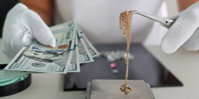 A jeweler weighs a gold chain on a precision scale while holding several banknotes in his other hand in a jewelry store